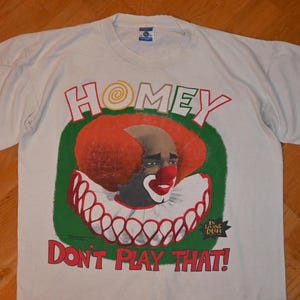 May include: A white short-sleeve t-shirt featuring a graphic of a clown with bright red hair and a red nose. The text above reads HOMEY and the text below reads DON'T PLAY THAT! with a small IN LIVING COLOR logo in the bottom right corner.