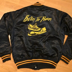 May include: A black satin bomber jacket with yellow and black striped cuffs and waistband. The back of the jacket features a yellow graphic of a pair of sneakers and the text "Born To Run".