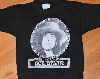 May include: Black t-shirt featuring a portrait of Bob Dylan in a hat, surrounded by a decorative border. The name "BOB DYLAN" is printed in a rectangular frame below the portrait. The shirt is made of a soft material.
