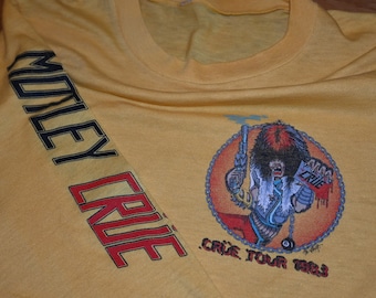 May include: Yellow long-sleeve t-shirt with a black and red "Motley Crue" graphic on the left sleeve and a cartoon illustration of a man with a gun and a chain on the right side. The illustration is in a circle with the text "Crue Tour 1983" underneath.