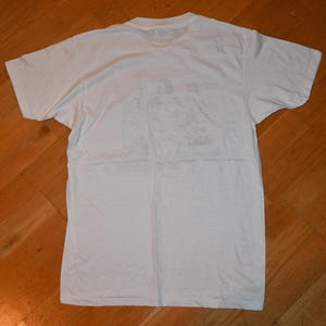 May include: A plain white short-sleeved t-shirt with a crew neckline. The garment is laid flat and features a faint, faded graphic print on the center of the chest area, showing indistinct figures and text that are not clearly legible.