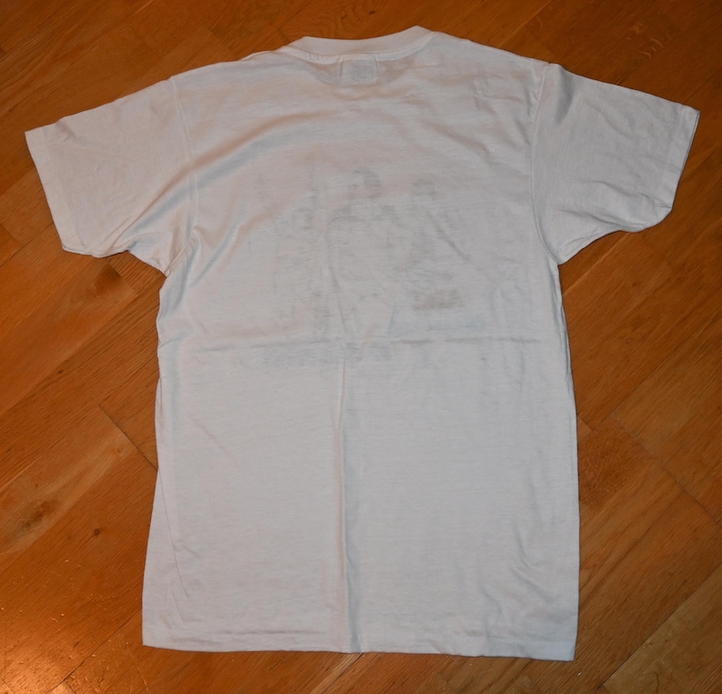 May include: A plain white short-sleeved t-shirt with a crew neckline. The garment is laid flat and features a faint, faded graphic print on the center of the chest area, showing indistinct figures and text that are not clearly legible.