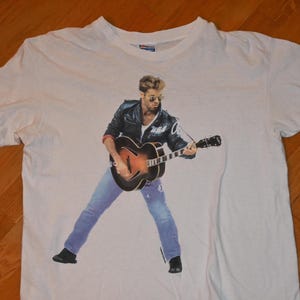 May include: White t-shirt featuring a graphic of a musician playing a guitar. The musician is wearing a black leather jacket, blue jeans, and sunglasses. The guitar is brown and black.