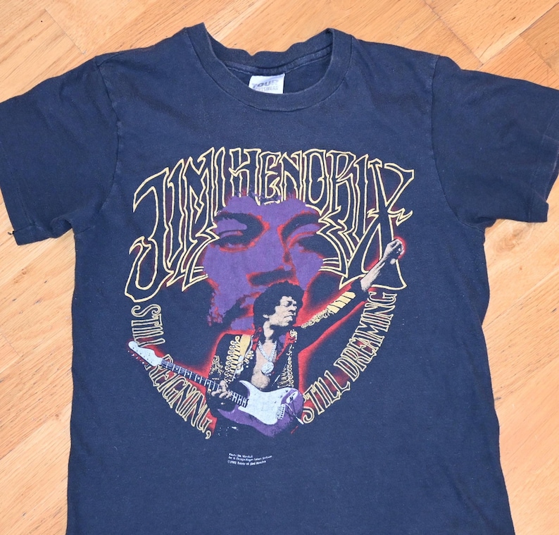 May include: Dark blue t-shirt with a graphic of Jimi Hendrix, featuring the text "Jimi Hendrix Still Dreaming". The design includes a purple face and a figure playing a guitar. The shirt has a crew neck.