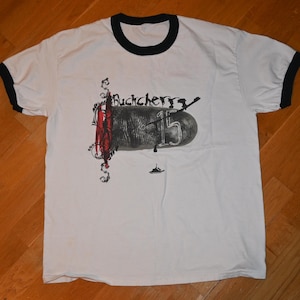 Könnte beinhalten: Weißes T-Shirt mit schwarzem Kragen und Bündchen. Auf der Vorderseite befindet sich der Bandname "Buckcherry" und die Zahl "15" in einem stilisierten Design, zusammen mit weiteren grafischen Elementen.