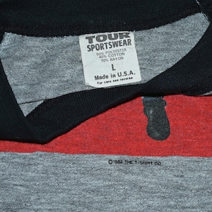 May include: Close-up of a vintage gray T-shirt with a black collar and red stripe. The shirt has a tag that reads "TOUR SPORTSWEAR" and "Made in U.S.A." The shirt is a size L and is made of polyester, cotton, and rayon. The copyright reads "&copy;1984 THE T-SHIRT CO."