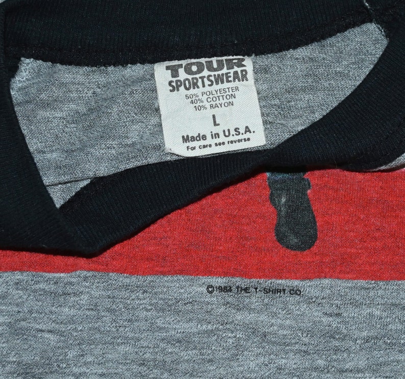 May include: Close-up of a vintage gray T-shirt with a black collar and red stripe. The shirt has a tag that reads "TOUR SPORTSWEAR" and "Made in U.S.A." The shirt is a size L and is made of polyester, cotton, and rayon. The copyright reads "&copy;1984 THE T-SHIRT CO."