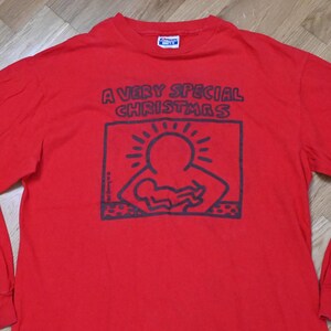 Könnte beinhalten: Rotes Langarm-T-Shirt mit einer schwarzen Grafik eines Strichmännchens, das ein Baby hält. Die Grafik befindet sich in einem weißen Kasten mit dem Text "A Very Special Christmas" darüber.