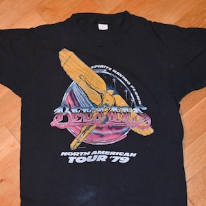 Puede incluir: Camiseta negra con un diseño gráfico que presenta el texto "Spirits Having Flown" y "Bee Gees North American Tour '79". El diseño incluye una imagen estilizada de una figura alada en amarillo y rosa.