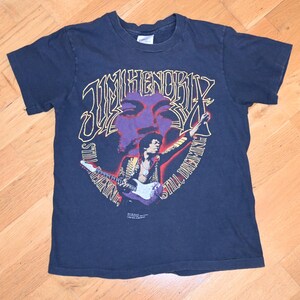 May include: Dark blue t-shirt with a graphic of Jimi Hendrix, the text "Jimi Hendrix" and "Still Dreaming". The graphic includes a purple face and a picture of Hendrix playing a guitar.
