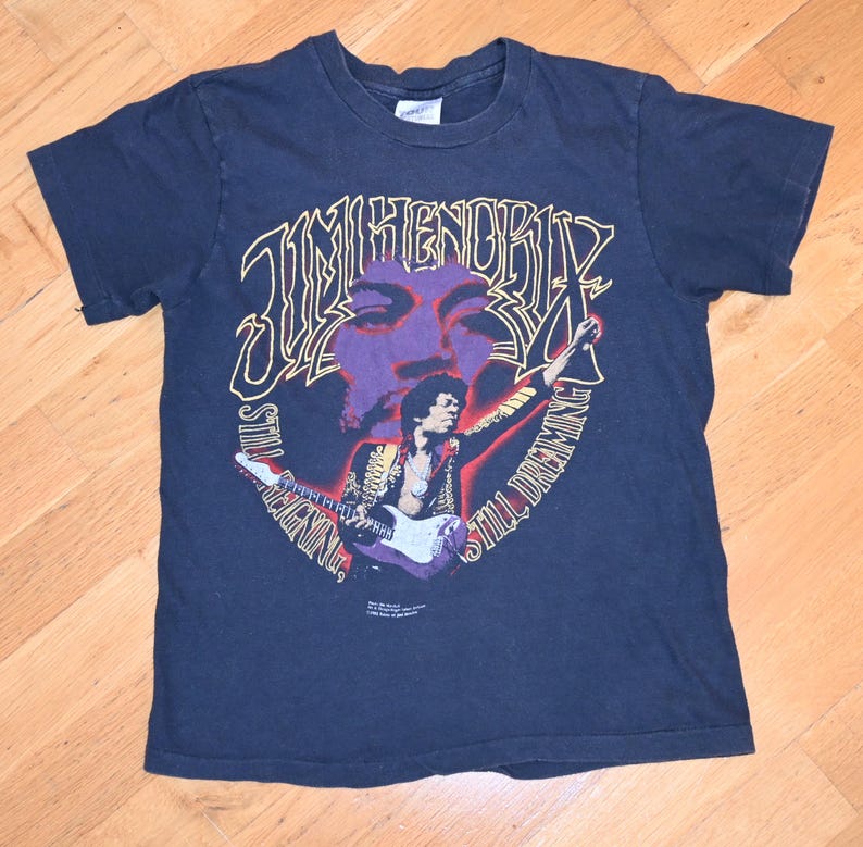 May include: Dark blue t-shirt with a graphic of Jimi Hendrix, the text "Jimi Hendrix" and "Still Dreaming". The graphic includes a purple face and a picture of Hendrix playing a guitar.