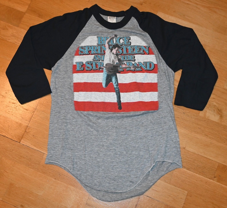May include: Gray raglan-style t-shirt with black sleeves. The front features a graphic of Bruce Springsteen and the E Street Band, with a photo of Springsteen in front of a red and white striped background.