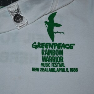 May include: White t-shirt with a green print of a bird, the words "GREENPEACE", "RAINBOW WARRIOR", "MUSIC FESTIVAL", and "NEW ZEALAND, APRIL 5, 1986".
