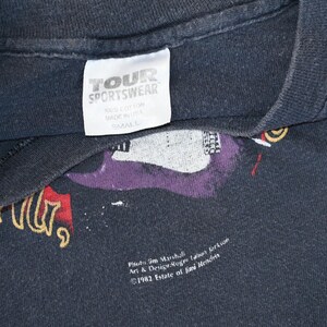 May include: Black vintage t-shirt with a graphic design featuring a purple guitar and text. The tag reads "TOUR SPORTSWEAR", "100% COTTON", "MADE IN USA", and "SMALL". The design includes the text "Photo Jim Marshall" and "&copy;1982 Estate of Jimi Hendrix".