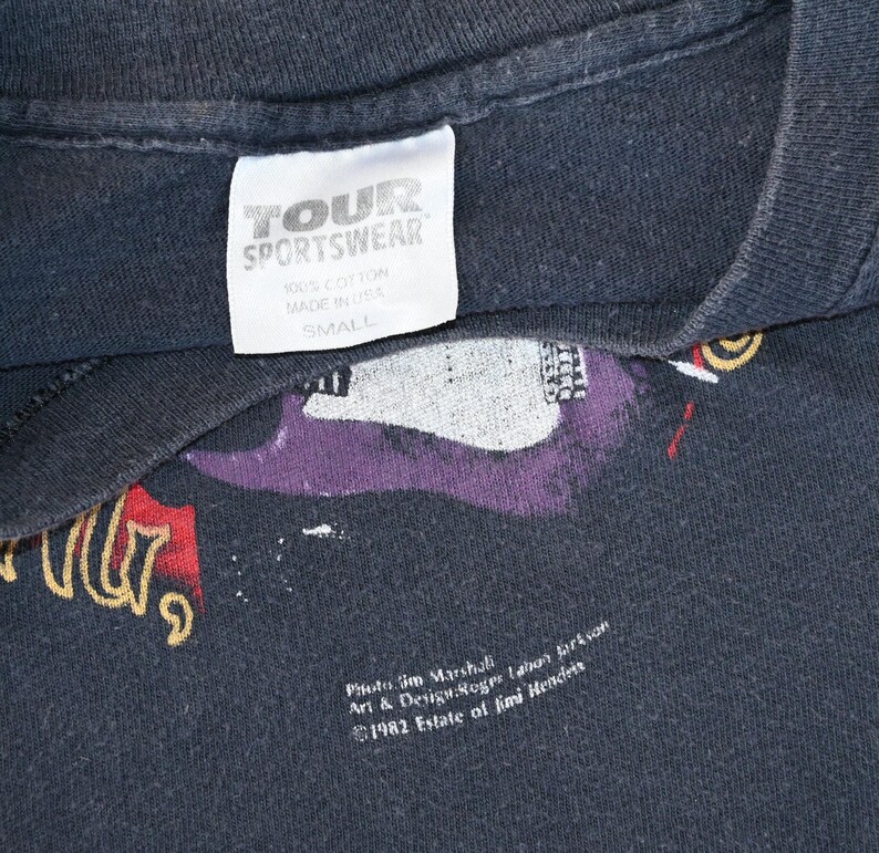 May include: Black vintage t-shirt with a graphic design featuring a purple guitar and text. The tag reads "TOUR SPORTSWEAR", "100% COTTON", "MADE IN USA", and "SMALL". The design includes the text "Photo Jim Marshall" and "&copy;1982 Estate of Jimi Hendrix".