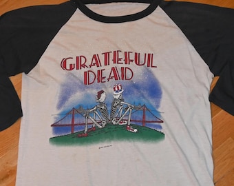 May include: A vintage baseball-style t-shirt with a cream-colored body and black sleeves. The shirt features the words "Grateful Dead" in red above an illustration of two skeletons, the Golden Gate Bridge, and a blue sky.