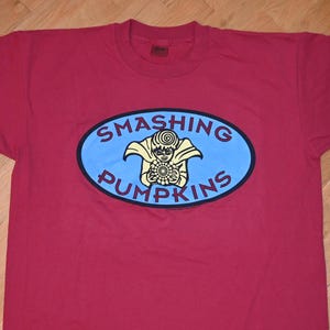 Puede incluir: Camiseta roja con un logotipo ovalado azul con el texto "Smashing Pumpkins" y un personaje de dibujos animados que sostiene un objeto giratorio.