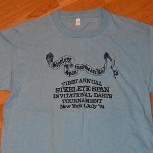 以下が含まれることがあります： 薄い青色のTシャツに、黒い文字で「Steeleye Span Now We Are Six」と「First Annual Steeleye Span Invitational Darts Tournament New York 1 July '74」と書かれています。