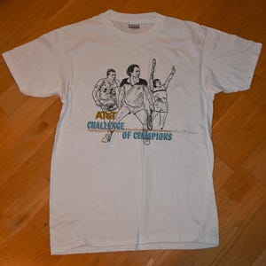 May include: A white short-sleeve t-shirt featuring a black line-art graphic of three tennis players in action. Below the figures, the text reads AT&T CHALLENGE OF CHAMPIONS in gold and blue lettering. The shirt has a crew neckline and a standard fit.