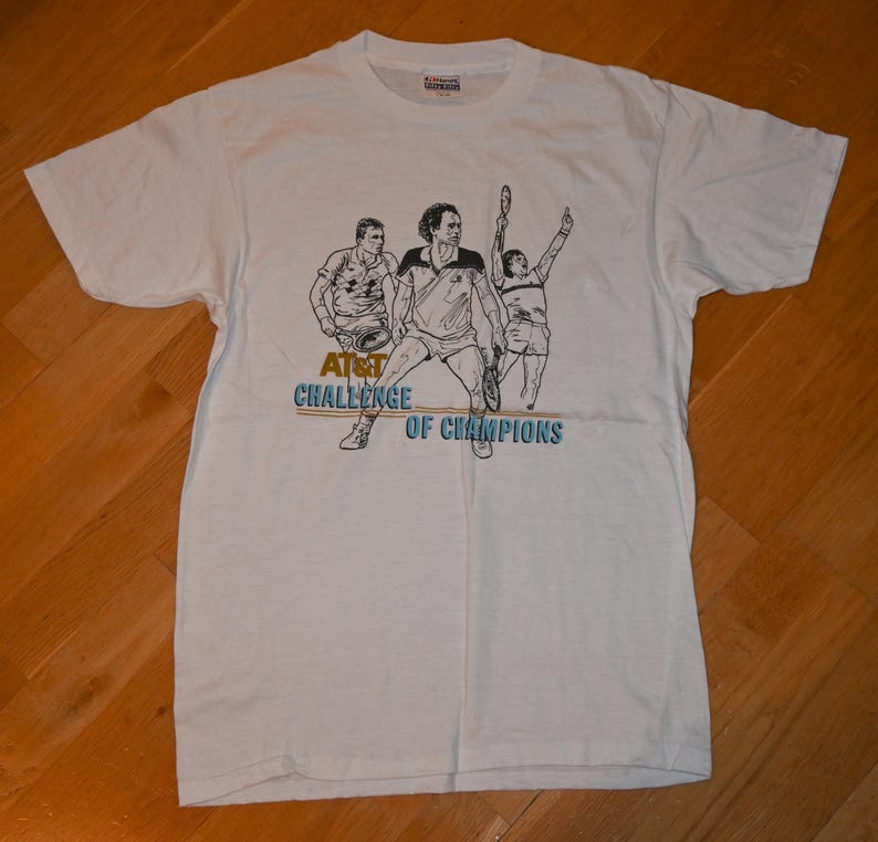 May include: A white short-sleeve t-shirt featuring a black line-art graphic of three tennis players in action. Below the figures, the text reads AT&T CHALLENGE OF CHAMPIONS in gold and blue lettering. The shirt has a crew neckline and a standard fit.
