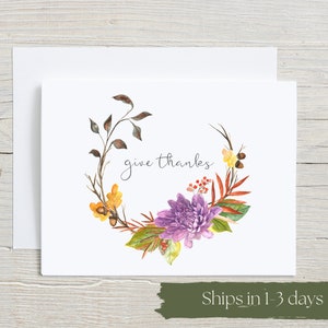 Give Thanks watercolor card with elegant script lettering surrounded by autumn florals. Purple chrysanthemums, golden flowers, fall leaves and berries create beautiful botanical wreath. Thanksgiving gratitude greeting card on white background with envelope on rustic wood.