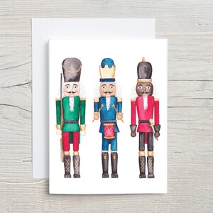 May include: Three watercolor nutcracker figurines. The nutcracker on the left is wearing a green uniform with a red sash and a blue hat. The nutcracker in the middle is wearing a blue uniform with a red sash and a blue hat. The nutcracker on the right is wearing a red uniform with a white sash and a blue hat.