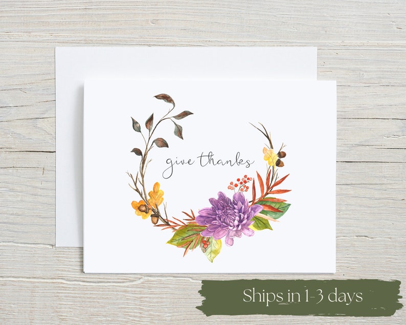 Give Thanks watercolor card with elegant script lettering surrounded by autumn florals. Purple chrysanthemums, golden flowers, fall leaves and berries create beautiful botanical wreath. Thanksgiving gratitude greeting card on white background with envelope on rustic wood.