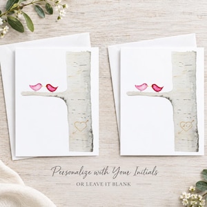 May include: Two white greeting cards featuring a watercolor illustration of a birch tree trunk with a branch. Two small pink birds sit on the branch. The tree trunk displays a gold heart outline with an arrow, one blank and one with initials.