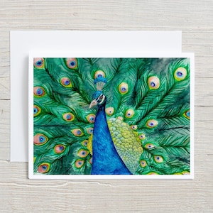 May include: A watercolor painting of a peacock with its feathers fanned out. The peacock's body is blue, with a crest of feathers on its head. The feathers are green with blue and yellow eye-like markings. The painting is on a white card.