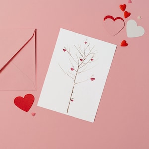 Puede incluir: Una tarjeta blanca con un dibujo de un &aacute;rbol desnudo con corazones rojos colgando de sus ramas. La tarjeta est&aacute; sobre una superficie rosa con un sobre rosa y corazones rojos esparcidos alrededor.