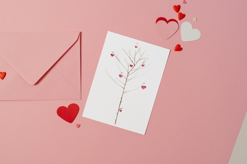 Puede incluir: Una tarjeta blanca con un dibujo de un &aacute;rbol desnudo con corazones rojos colgando de sus ramas. La tarjeta est&aacute; sobre una superficie rosa con un sobre rosa y corazones rojos esparcidos alrededor.