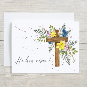 May include: A white card with a wooden cross surrounded by watercolor flowers. The text "He has risen!" is printed below the cross.