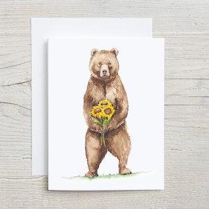 May include: A watercolor illustration of a brown bear holding a bouquet of yellow sunflowers. The bear is standing on a green patch of grass.