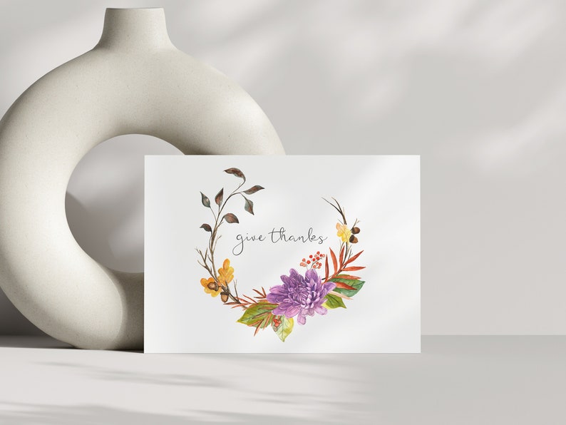 May include: A white card with a watercolor illustration of a floral wreath with purple flowers, yellow leaves, and brown branches. The text "give thanks" is written in cursive on the card.