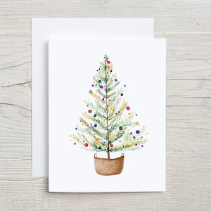 May include: A watercolor painting of a Christmas tree with colorful ornaments. The tree is in a brown wicker basket and is on a white background.