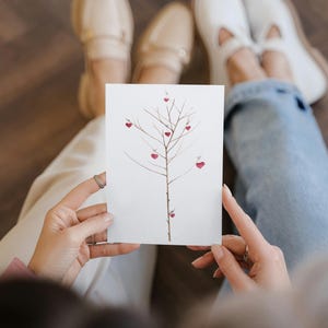 Puede incluir: Una tarjeta blanca con una pintura de acuarela de un &aacute;rbol con corazones rojos en las ramas. La tarjeta est&aacute; siendo sostenida por dos personas.