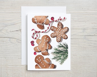 May include: Watercolor Christmas card featuring gingerbread men and a snowflake cookie with white icing. The card also includes red berries, a red and white striped string, and a sprig of evergreen. The card is on a white background.