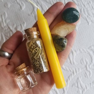 May include: A hand holding a yellow candle, a small glass bottle with dried herbs, a clear glass bottle, and three stones: a green stone, a light green stone, and a clear stone.
