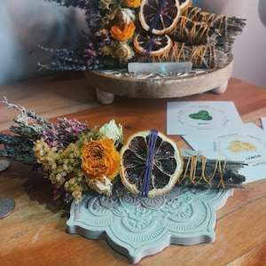 May include: Two bundles of dried herbs and flowers with dried citrus slices. The bundles are tied with twine and are sitting on a decorative wooden and a concrete base. The bundles are labeled "Lemon" and "Protection, Prosperity, Abundance, The Money Plant, Success".