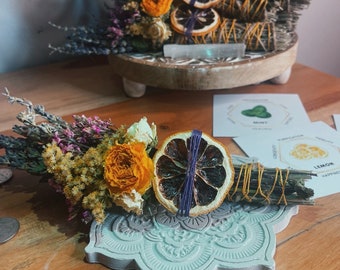 May include: Two bundles of dried herbs and flowers with dried citrus slices. The bundles are tied with twine and are sitting on a decorative wooden and a concrete base. The bundles are labeled "Lemon" and "Protection, Prosperity, Abundance, The Money Plant, Success".