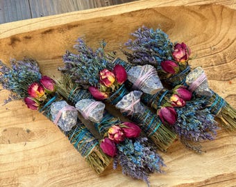 Op de afbeelding: Vier bundels gedroogde kruiden omwikkeld met blauw touw, elk versierd met kleine roze rozenknopjes, takjes blauwe statice en een grijze steen vastgezet met dun roze draad, allemaal samen gerangschikt in een lichtbruine houten kom.
