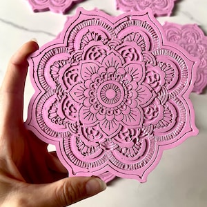 Pode incluir: Quatro  tapetes de copo de resina rosa com um design de mandala detalhado. Os tapetes de copo têm a forma de uma flor com múltiplas camadas de padrões intrincados.