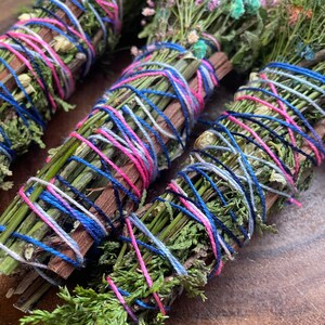 May include: Three bundles of dried herbs and flowers tied together with pink, blue, and white yarn. The bundles are arranged on a wooden surface.