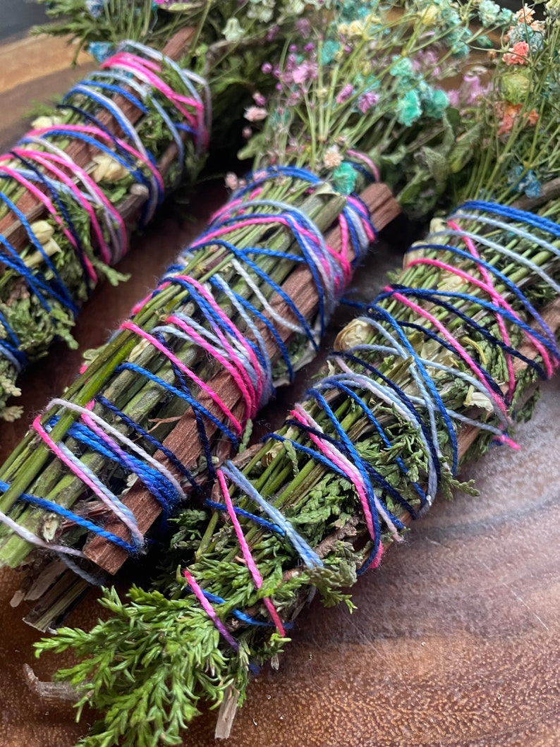 May include: Three bundles of dried herbs and flowers tied together with pink, blue, and white yarn. The bundles are arranged on a wooden surface.