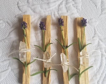 May include: Four wooden sticks with lavender flowers and clear quartz crystals tied to them. The sticks are on a white fabric background.