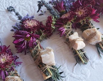 May include: Bundles of dried flowers and herbs, including purple chrysanthemums, lavender, and green herbs, are tied with twine and feature a light-colored stone. The bundles are arranged on a white, textured surface.