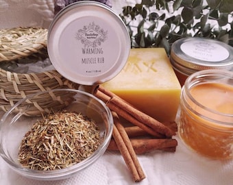 May include: A silver tin of warming muscle rub with a label that reads "Charmed Herbs" and "Warming Muscle Rub" with a 4 oz / 118 ml size. The tin is surrounded by cinnamon sticks, dried herbs, a bar of soap, and a jar of honey.