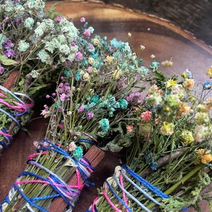 May include: Three bundles of dried flowers and herbs tied together with colorful string. The bundles are made with a variety of dried flowers, including white, pink, blue, and yellow flowers. The bundles are tied with blue, pink, and white string.