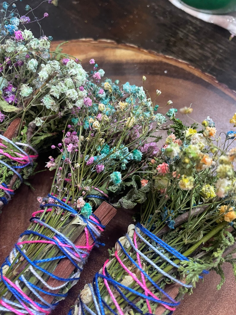 May include: Three bundles of dried flowers and herbs tied together with colorful string. The bundles are made with a variety of dried flowers, including white, pink, blue, and yellow flowers. The bundles are tied with blue, pink, and white string.