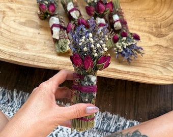 Op de afbeelding: Een bundel gedroogde groene dennennaalden vastgebonden met magentakleurig draad, voorzien van een bosje gedroogde paarse lavendel, wit gipskruid en donkerroze rozenknoppen, met een kleine grijs-wit gespikkelde steen aan de zijkant bevestigd.
